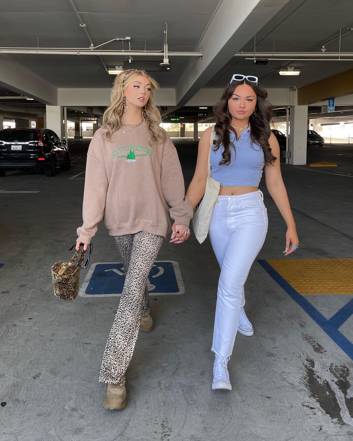 two young women are walking handinhand towards the camera in an indoor parking garage. the woman on the left has blonde hair and is wearing a tan sweatshirt with a graphic, leopard print pants, and tan shoes. she carries a leopard print purse. the woman on the right has brunette hair and is wearing a light blue cropped polo shirt, white jeans, and white sneakers. she carries a white tote bag and has sunglasses perched on her head.