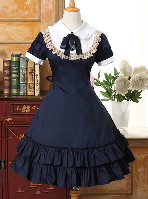 a navy blue and white lolita fashion dress is displayed on a mannequin. the dress features a white collar with lace trim, a black bow at the neck, and a tiered, ruffled skirt. the background includes a wooden cabinet with books and a white vase with flowers.