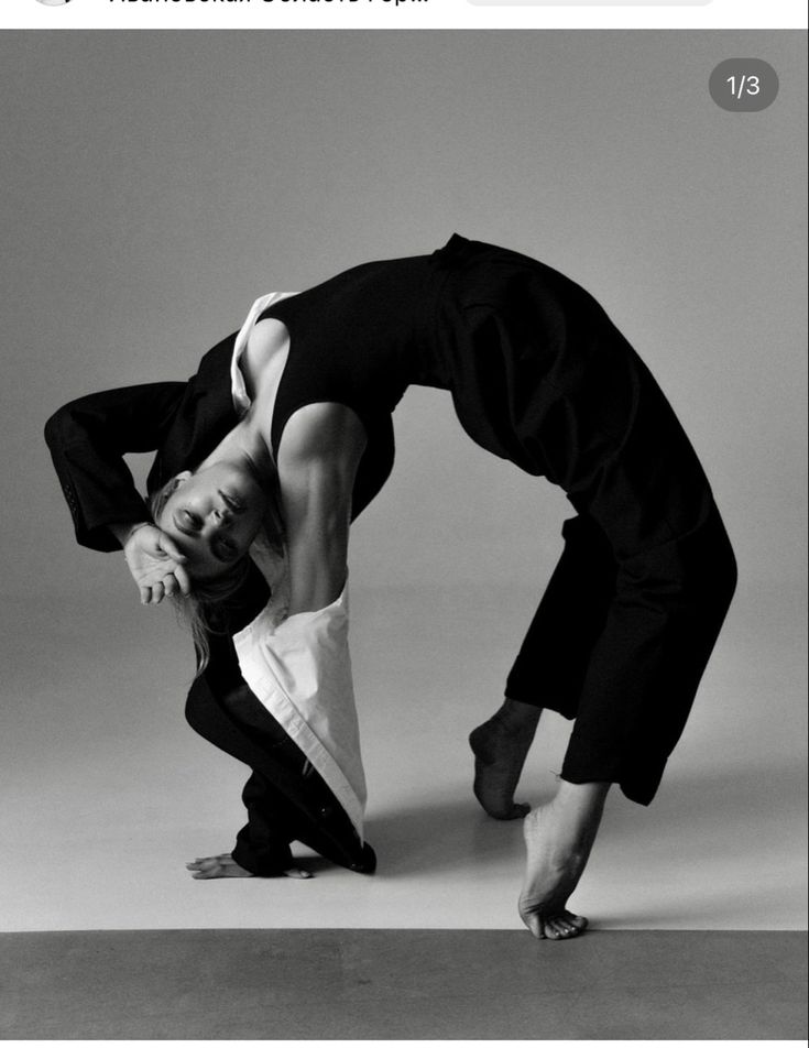 a black and white photograph depicts a woman, seemingly a dancer, performing a graceful backward bend. she is wearing a black jumpsuit that resembles a pantsuit, with a white shirt draped over her shoulders. her body is arched, showcasing impressive flexibility and athleticism. the image is taken in a studio setting on a plain floor, highlighting the dancer and her pose. the overall style is artistic and elegant, emphasizing the dancer's strength and grace.