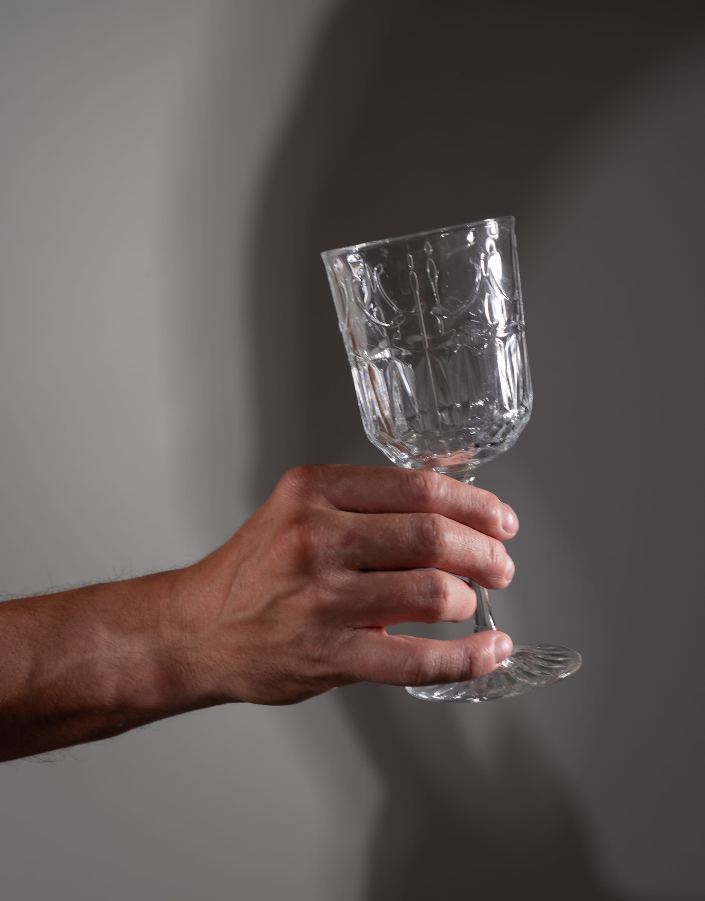 a man's hand holds a clear cut crystal wine glass up against a gray background. the image is lit with dramatic lighting that creates strong shadows of both the hand and the glass.