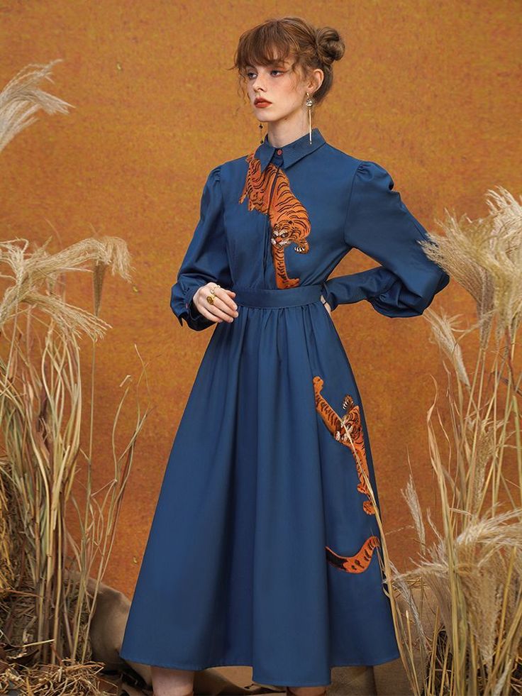 a fulllength studio shot depicts a woman wearing a stylish, dark blue midi dress. the dress features a collared shirt bodice with long sleeves and an aline skirt.  an orange tiger print is prominently featured on the shirt and skirt. the model is posing against an orange backdrop with tall dried grasses. she has her hair styled up and is wearing simple earrings. the overall style suggests a vintage or retro aesthetic.