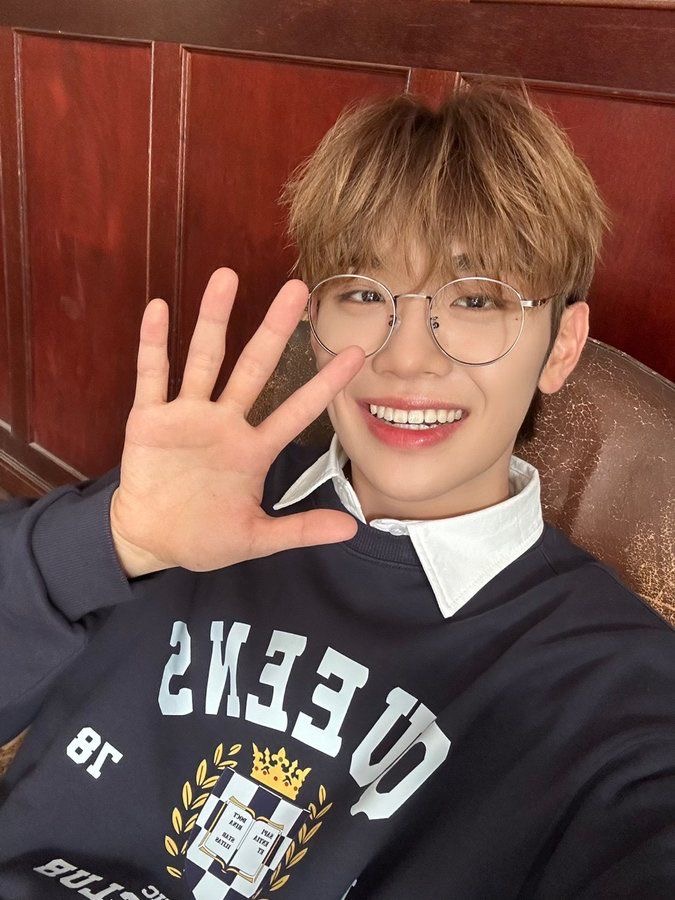a young man with bleached blonde hair and round glasses smiles brightly at the camera while taking a selfie. he holds up his left hand in a wave, revealing a silver ring on his pinky finger.  he's wearing a dark navy blue sweatshirt with a collegiatestyle graphic and a white collared shirt underneath. the background features a textured wall of warm reddishbrown wood paneling. the image exudes a friendly and approachable atmosphere.