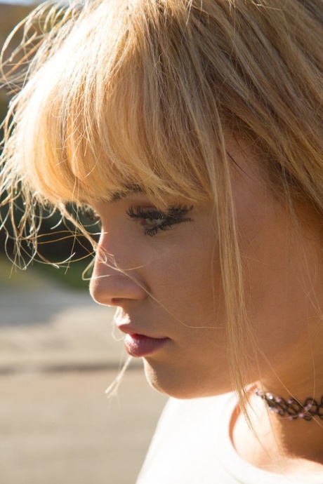 the image shows a woman with blonde hair styled in a messy, textured look with bangs. she is wearing a black choker necklace and a white shirt. the photo is a side profile shot, capturing the woman's face and neck. her expression is neutral, with her lips slightly parted. the lighting is natural and bright, casting shadows on her face and creating a warm tone. the background is blurred, suggesting an outdoor setting. the focus is on the woman's profile and the details of her features.