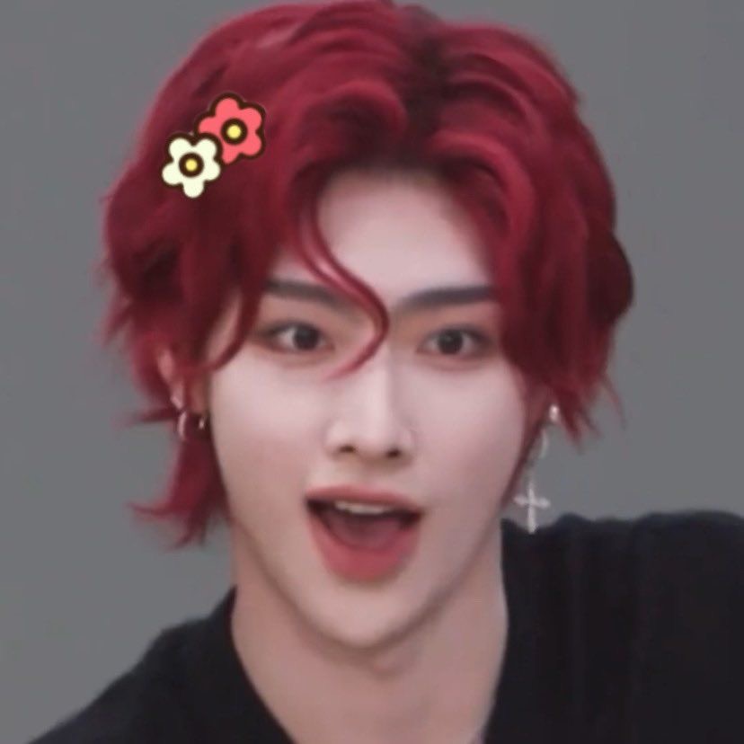 the image is a closeup portrait of a young asian man with vibrant red hair styled in a trendy, wavy fashion. he wears minimal makeup that accentuates his features, and his expression is a mix of surprise and amusement with his mouth slightly open.  the lighting suggests a studio environment, possibly for a photoshoot or performance. his overall style, from the hair to the subtle ear piercings, hints at a kpop idol aesthetic.