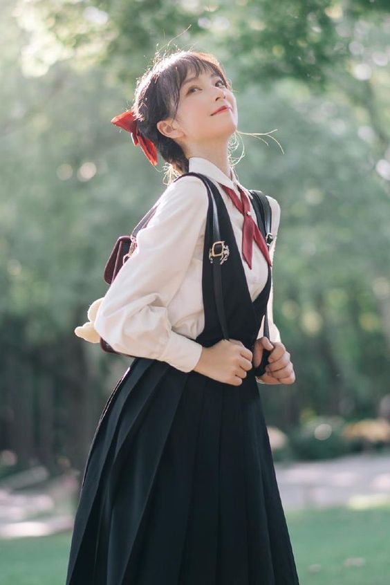 young woman, wearing a stylish black dress, white blouse, and red bow, stands in a park with her backpack on. the dress features suspenders and a pleated skirt. she is looking up with a cheerful expression, bathed in soft sunlight filtering through the trees. her outfit and pose suggest a trendy japanese fashion style, possibly inspired by lolita fashion.