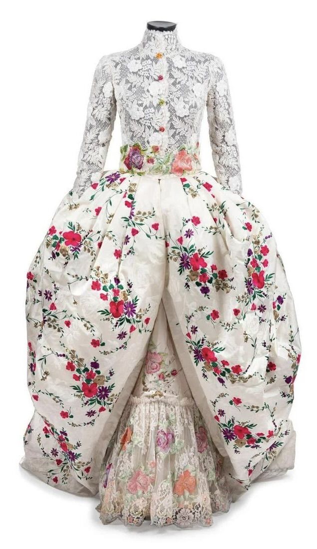 a formal gown is displayed on a mannequin. the top portion of the dress features long lace sleeves and a high collar, fastening with buttons. a floralpatterned belt cinches the waist. the skirt is full and flowing, adorned with a white base and a floral print. the lower section reveals a decorative layer with intricate floral embroidery and lace detailing.