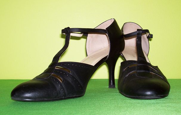 two black tstrap heeled shoes are displayed against a green and yellow background. the shoes are leather with a closed toe and a delicate strap that crosses the foot and buckles at the ankle. the heels are slim and elegant.