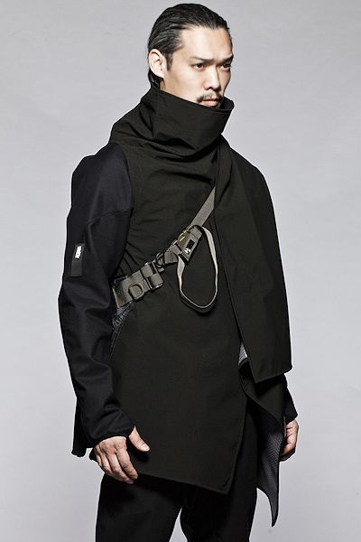 a male model stands in a studio setting, showcasing a contemporary black jacket with an asymmetrical design and a high collar. the jacket features utilitarian elements such as straps and buckles.  the model's pose and the garment's style convey a sense of urban edge.