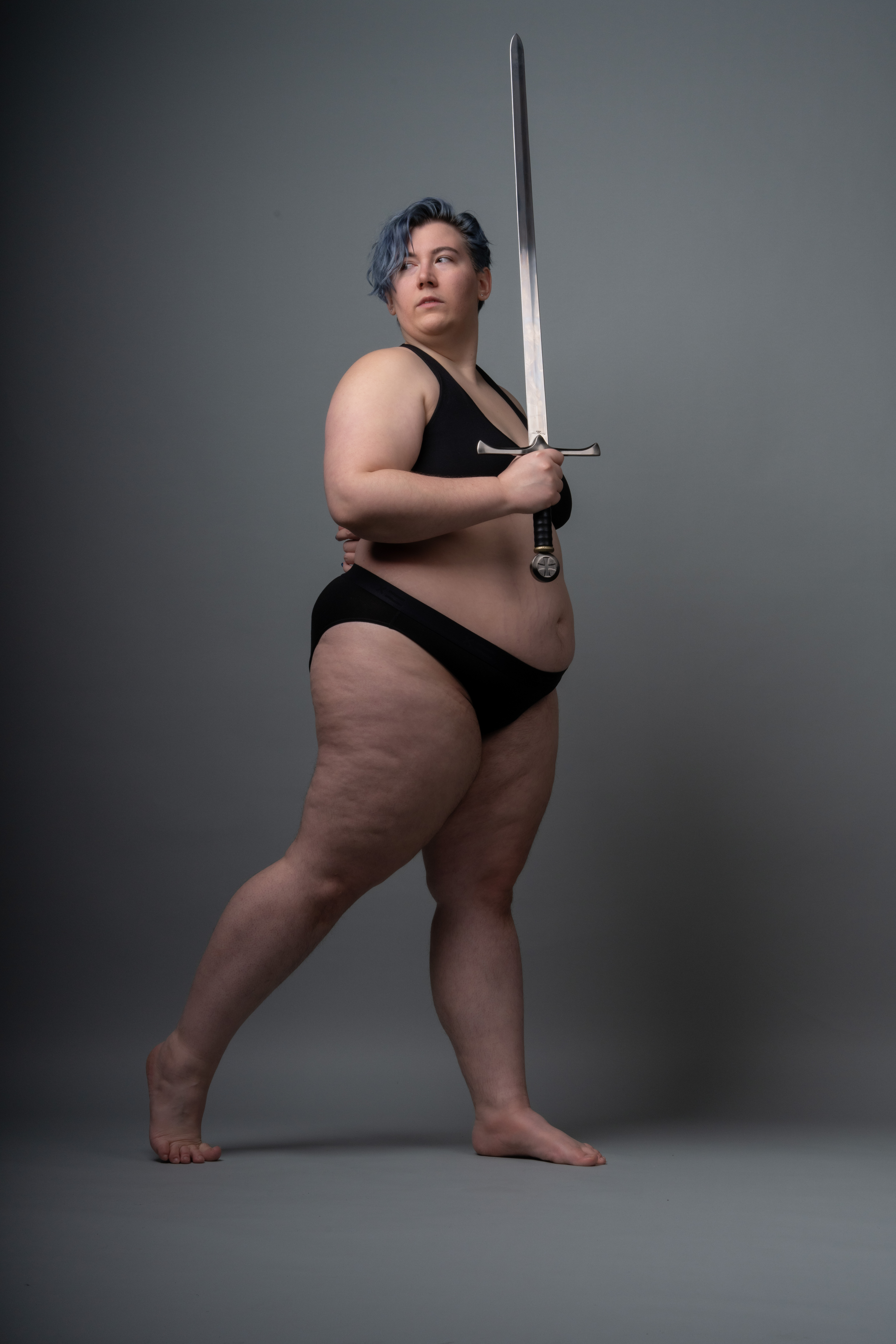a fulllength studio shot of a woman with blue hair standing barefoot on a grey background, holding a sword. she is wearing black underwear. she is looking off to the side.