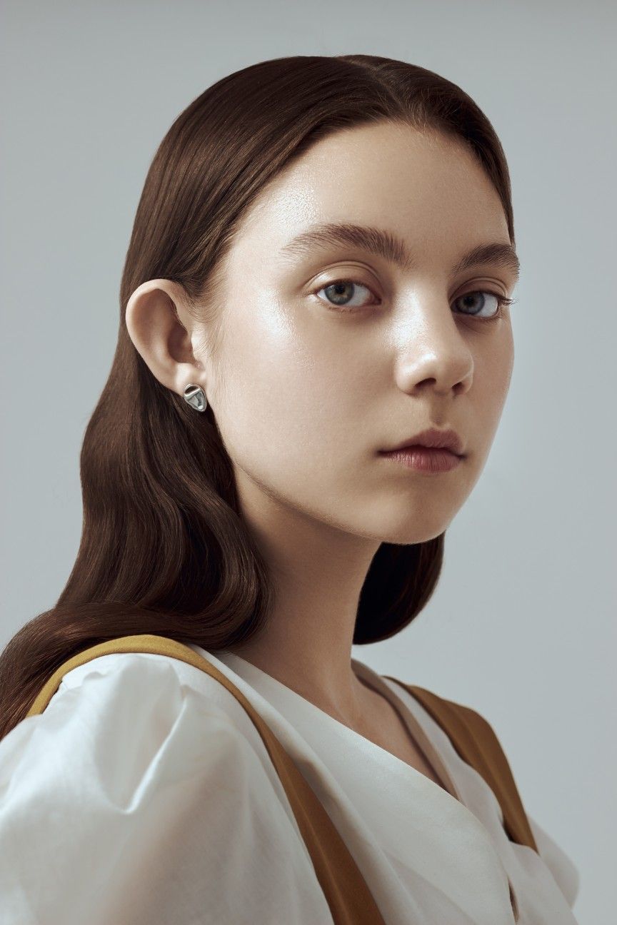 closeup portrait of a young woman with long brown wavy hair. she is wearing a white blouse with a subtle mustardcolored strap detail.  her makeup is minimal, highlighting her clear complexion. she has a serious expression and is looking slightly offcamera. the background is a plain, light gray. the overall mood is sophisticated and understated.
