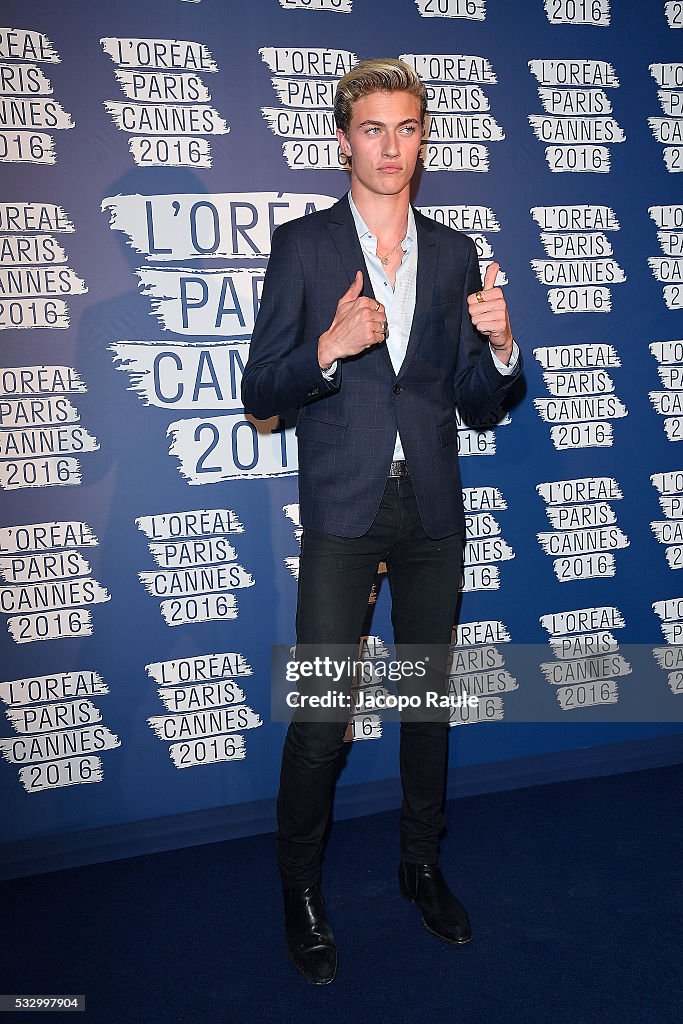 a man is standing in front of a l'oreal paris cannes 2016 backdrop. he is wearing a navy blue suit with a white collared shirt underneath, black pants, and black dress shoes. he is holding his hands up with his thumbs up.