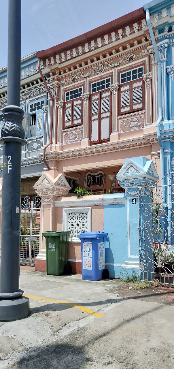 an old, colorful shophouse building in singapore with ornate details and windows. the building is on a street with a sidewalk and a streetlight. there are two trash cans in front of the building. the building is a mix of different colors, including blue, yellow, pink, and white. the windows are arched and have intricate designs. the building is in a good condition and wellmaintained. it is a typical example of the traditional shophouse architecture of singapore.  the building is in the background and the street scene in the foreground, with a streetlight and trash cans adding depth to the image.