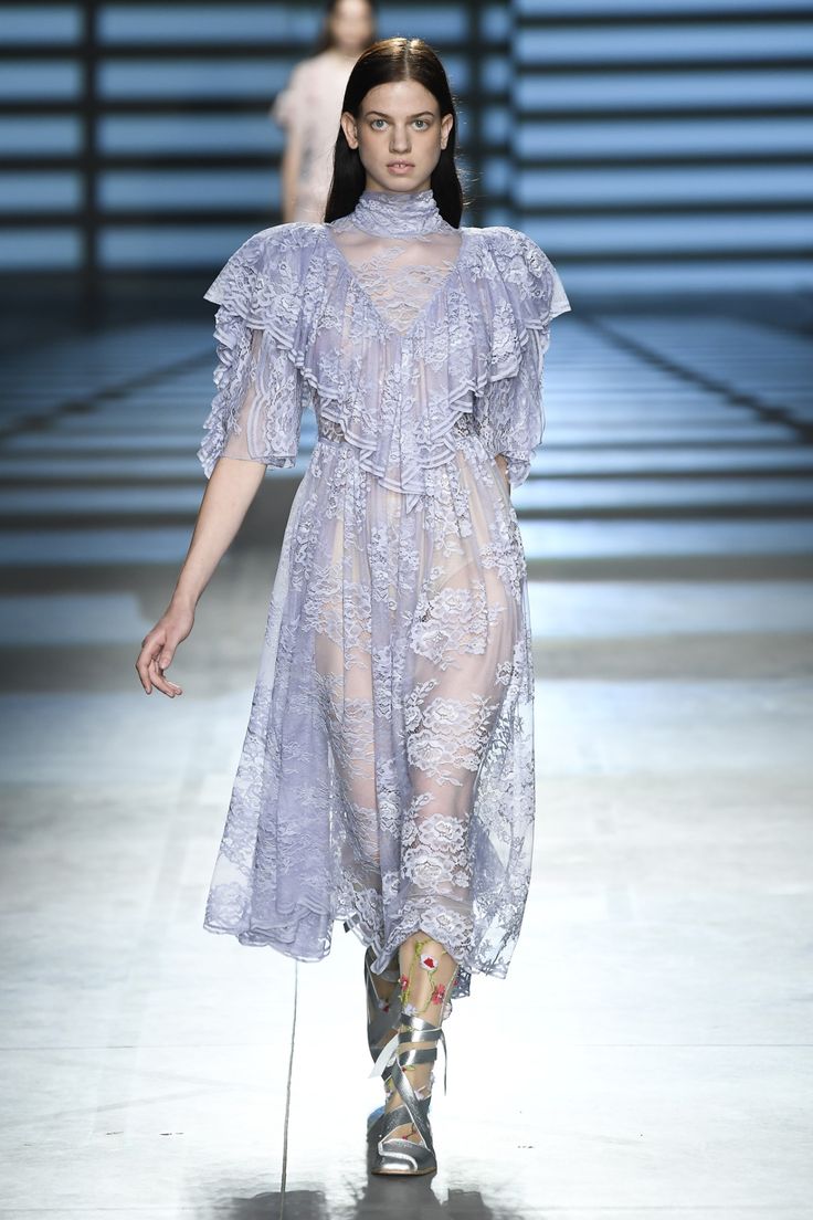 a model walks down a runway wearing a light lilac sheer lace dress. the dress has frills and ruffles at the shoulders and bodice.  the model is wearing highnecked, long sleeved dress and silver gladiatorstyle ankle boots with floral embroidery on the leg coverings.  the background is a blurred out modern setting.