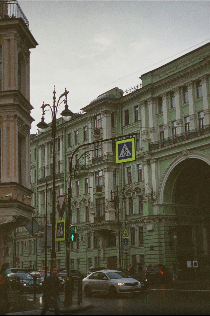 a moody, atmospheric photograph taken on a cloudy day in what appears to be st. petersburg, russia. the image showcases a picturesque street scene, with old, historical buildings lining the sides. the architecture is elaborate, with ornate details and pastel green facades.  cars are parked and driving along the street, which is slightly wet. streetlights and pedestrian crossing signs add to the urban atmosphere. the overall mood is tranquil yet bustling, captured with a filmlike quality.  the image focuses on the architectural details and urban scene with the cars as secondary subjects.
