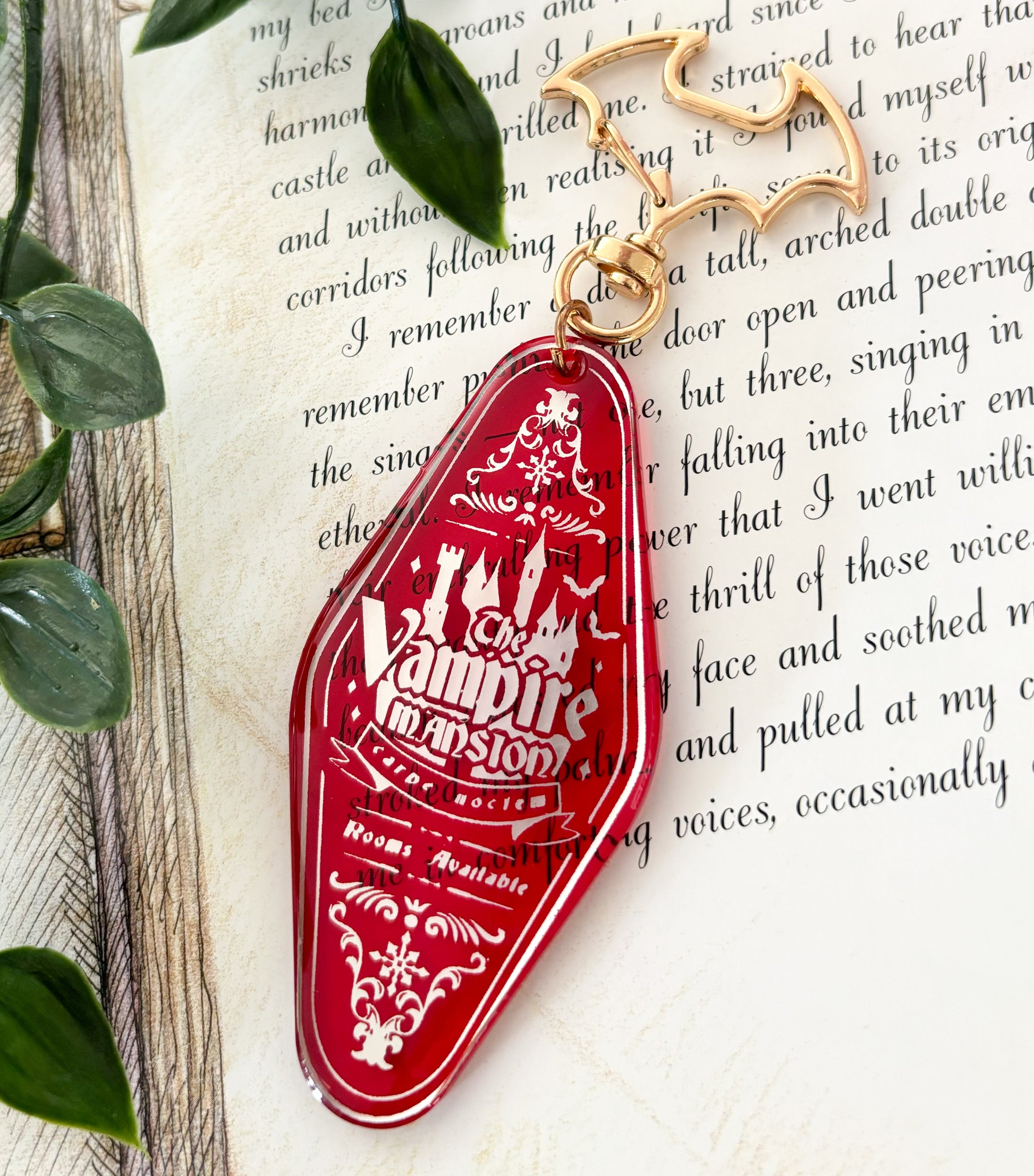 the image shows a closeup of a red acrylic keychain shaped like a vintage motel key. the keychain features a white engraved design that says "the vampire mansion" along with other decorative elements like bats and a castle.  it's resting on a page of a book with gothicstyle text. the keychain includes a goldcolored batshaped key ring. the background includes part of an open book with text, suggesting a literary or gothic theme. the overall aesthetic is spooky, vintage, and halloweenthemed.