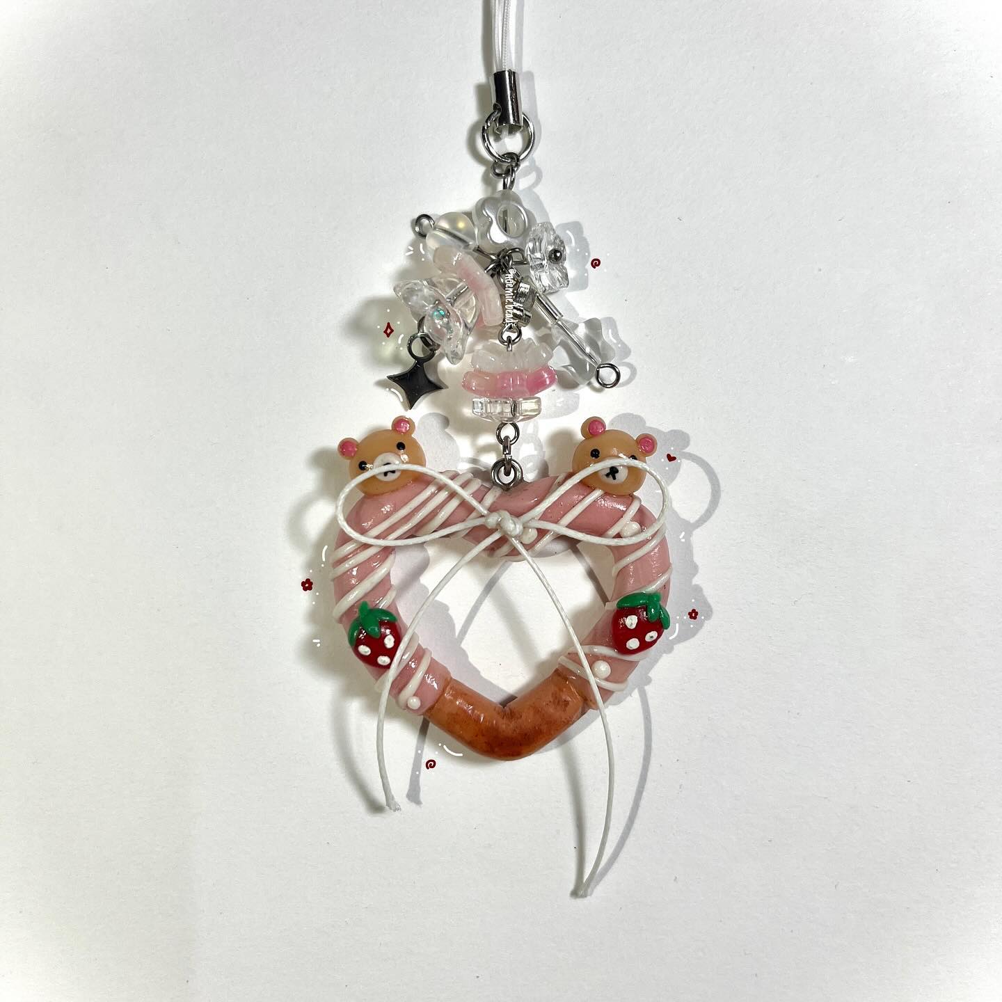 a handmade charm featuring a heartshaped base decorated with pink and white stripes, topped with miniature bears and strawberries. it has clear and plastic beads, a ribbon, and a silver keychain attachment.