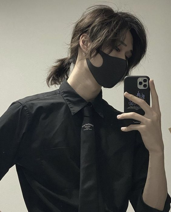 a young man with long, dark hair partially tied back is taking a mirror selfie. he is wearing a black buttondown shirt, black tie, and a black mask. the lighting is soft and artificial, and the overall aesthetic is minimalist and slightly androgynous.