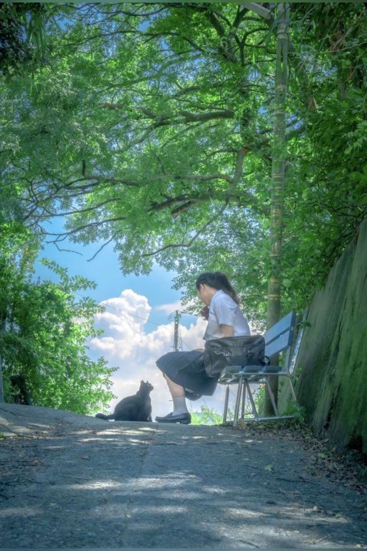 a woman is sitting on a blue bench in a park, with a black cat sitting nearby. the woman has dark hair pulled back in a ponytail and is wearing a white shirt and dark skirt. the cat is facing away from the woman, looking towards the sky. the bench is in a shady area with trees and greenery surrounding it. the sky is blue with fluffy white clouds. the image is taken from a low angle, looking up at the woman and cat.