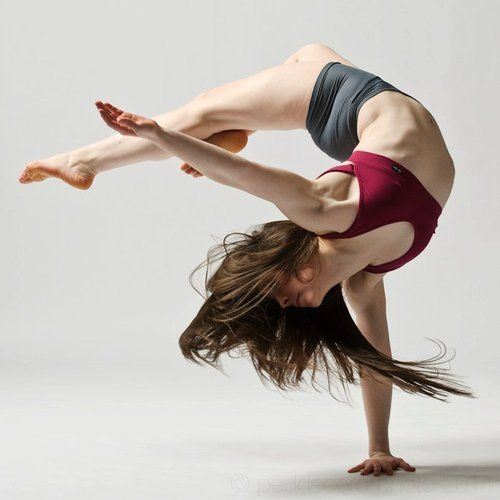 a female dancer is captured midair in a dynamic pose, showcasing her exceptional flexibility and strength. she balances on one hand, her body forming a graceful arc. the image is set in a bright, white studio, highlighting the dancer's form and movement. her long hair flows freely, adding to the sense of energy and motion.