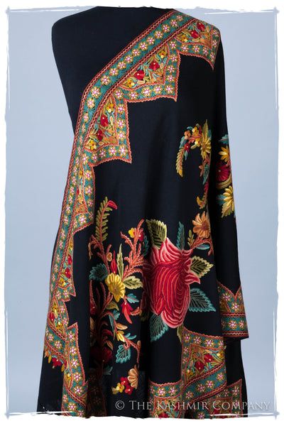 a black shawl with intricate floral embroidery is draped over a mannequin. the embroidery features a variety of flowers in red, yellow, orange, and green, with detailed borders of geometric and floral patterns. the shawl is from the kashmir company, as indicated by the watermark.