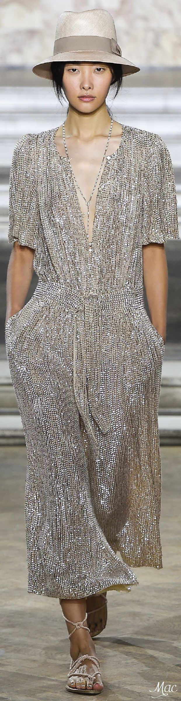 a female model is walking down a runway wearing a silver sequin dress, a widebrimmed hat, sandals, and jewelry. her dress is fitted and flows elegantly as she walks. she appears to be a fashion model in a runway show.