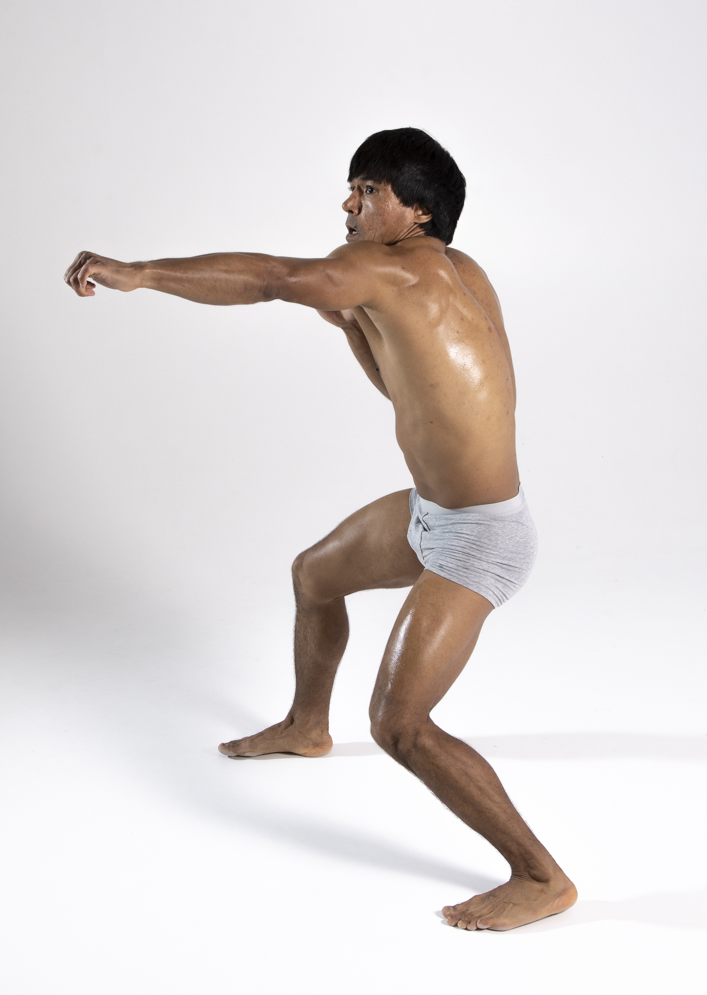 a barechested man wearing underwear is captured in a studio shot performing a martial arts move. he is positioned in a boxing stance, extending his arm forward as if punching. the man is visibly sweating, highlighting his athletic physique. the backdrop is a plain white background, emphasizing the man's figure and movements.