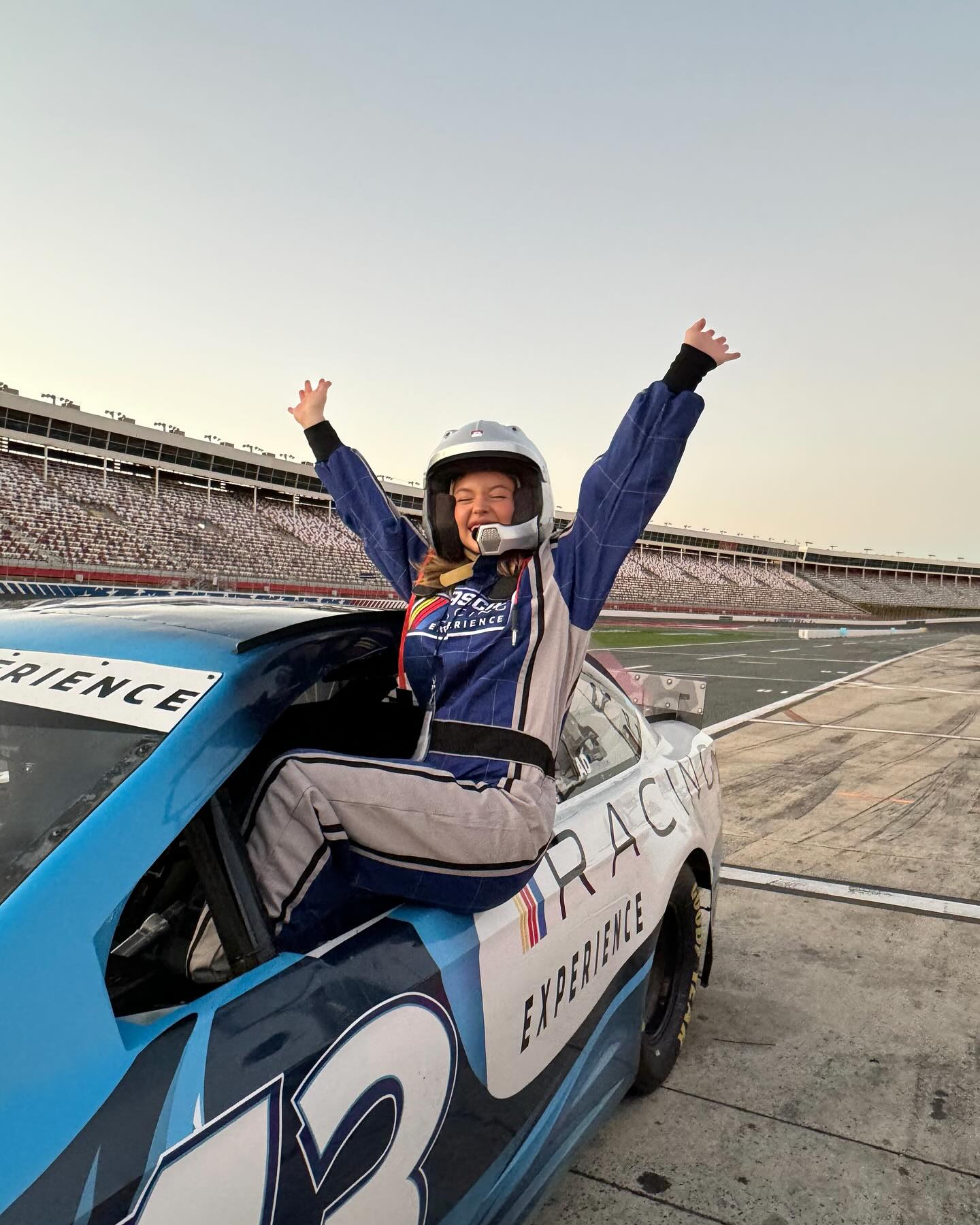 the image shows a young girl, possibly a teenager, wearing a racing helmet and suit, sitting triumphantly in a blue race car on a race track. her arms are raised in celebration. the car has the number 43 visible and branding that reads "racing experience."  the background shows a large grandstand with spectators and a clear view of the race track. the overall impression is one of joy and accomplishment, suggesting a successful racing experience. the lighting suggests the photo was likely taken at dusk or dawn.