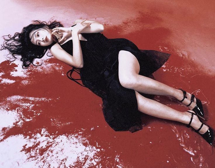 the image depicts a woman with long dark hair lying on a surface that appears to be stained red, possibly simulating blood. she wears a black dress and black highheeled shoes. her pose is languid and somewhat dramatic; she holds what appears to be a small object in her hands near her chest. the overall atmosphere of the picture is dark, moody, and slightly mysterious, suggestive of a fashion or film noirinspired photoshoot. the red background dominates the image, adding to the dramatic effect.