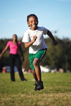 a young boy with a joyful expression is captured midstride, running across a grassy field on a bright sunny day. he's wearing a white tshirt, green athletic shorts, and running shoes. in the background, a woman in pink and black athletic wear is also present, possibly a parent or guardian. the overall atmosphere exudes happiness, energy, and the freedom of outdoor play.