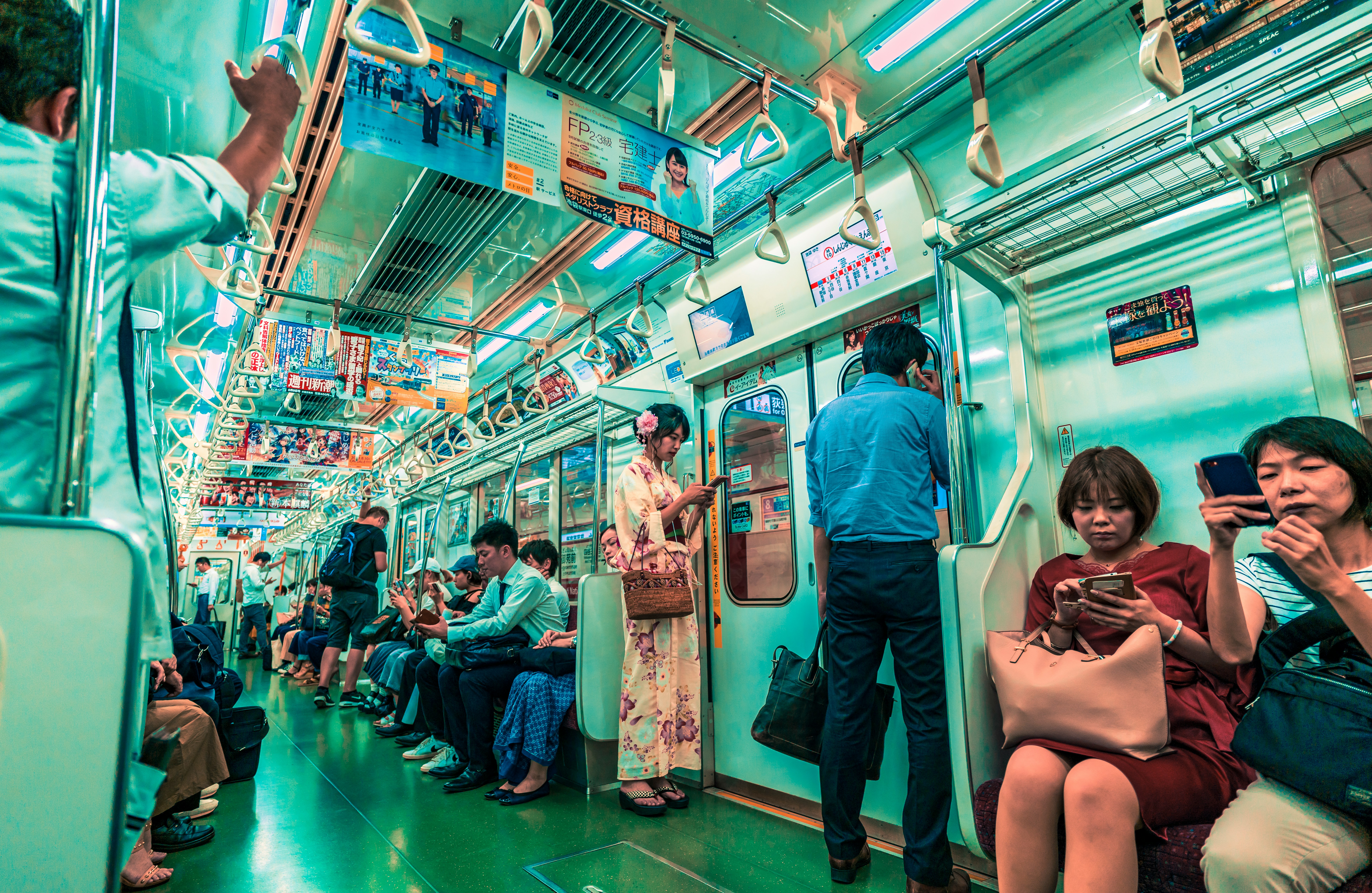 the image shows a view inside a crowded train car in japan. passengers are sitting and standing, many using their smartphones. various advertisements adorn the walls and overhead spaces of the train. a woman in a yukata stands near the train doors, while other passengers are seated, looking at their phones. the overall ambiance is urban, capturing a moment of daily commute. the color palette is predominantly teal.