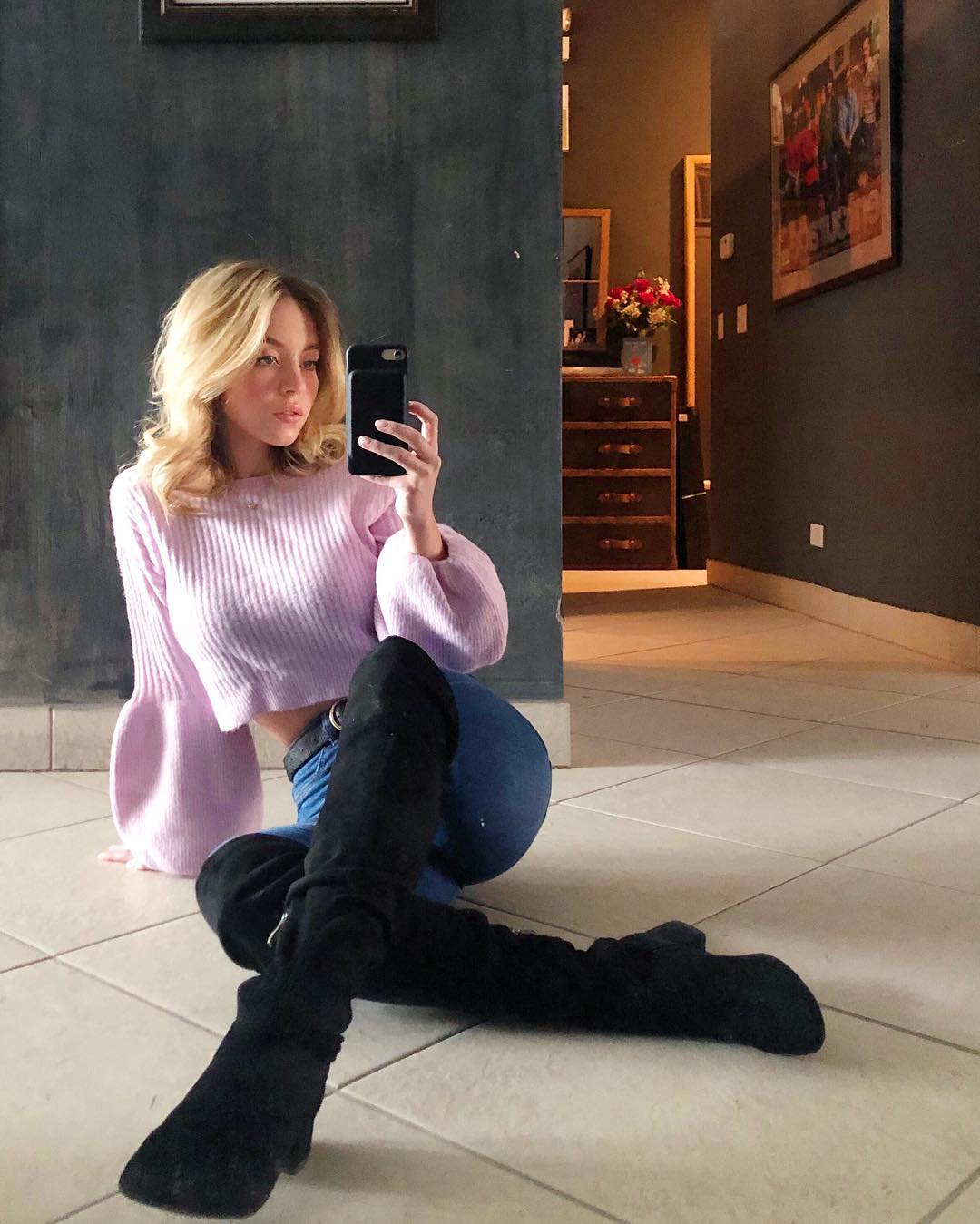 a young blonde woman is sitting on a tiled floor in a home, taking a selfie. she's wearing a pale pink cropped sweater with balloon sleeves, dark blue jeans, and tall black overtheknee boots.  the background shows a glimpse of a hallway with furniture, and a framed picture on the wall. the overall mood is casual and relaxed.