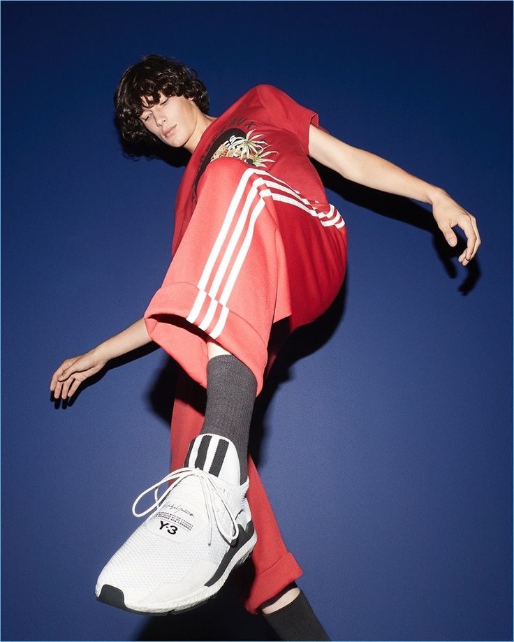 a young man in a red adidas tracksuit is posing in a studio with a blue background. he's wearing a white y3 sneaker with black stripes and socks. his hair is black and he's looking down at the camera.