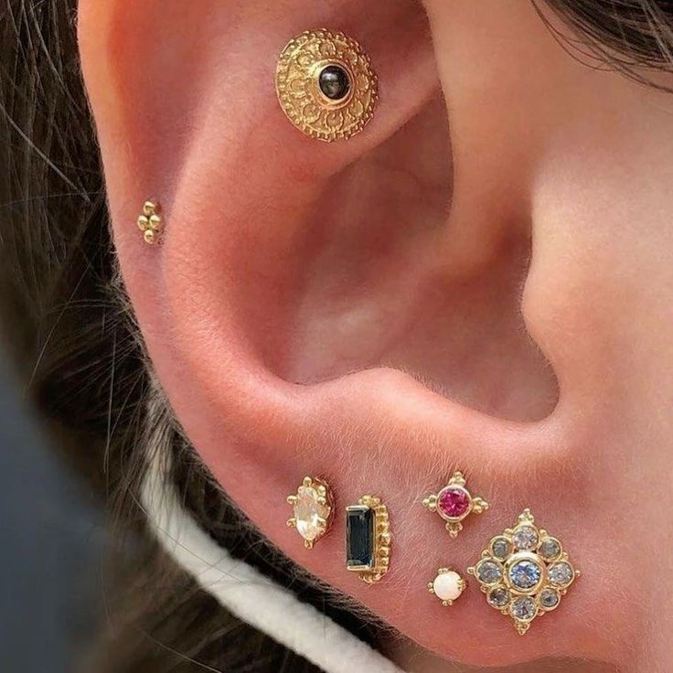 the image is a closeup shot of a person's ear, showcasing a variety of small, delicate gold piercings.  the piercings include a larger ornate gold disc with a dark stone at the top of the ear, a small cluster of three gold balls slightly lower.  the lower part of the ear features several smaller studs set in gold, including pieces with opal, black stone, ruby and a sapphire. the overall style of the piercings is dainty and feminine, with intricate detailing on several of the studs.  the ear is welllit, and the image has a high resolution making the details of the jewelry easily visible.
