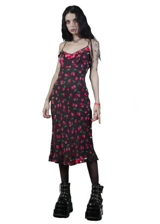 a fullbody studio shot of a young woman with dark hair and tattoos modeling a darkcolored midi dress with a cherry print. the dress features thin straps and a ruffle hem. she is wearing black platform boots and minimal accessories. the background is plain white. the overall style is dark, gothic, or alternative.