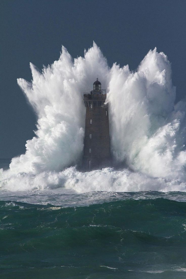 a tall, grey lighthouse is surrounded by a massive wave crashing against it. the wave creates a dramatic spray of white foam that nearly engulfs the structure. the ocean water is deep green and choppy, and the sky is overcast.