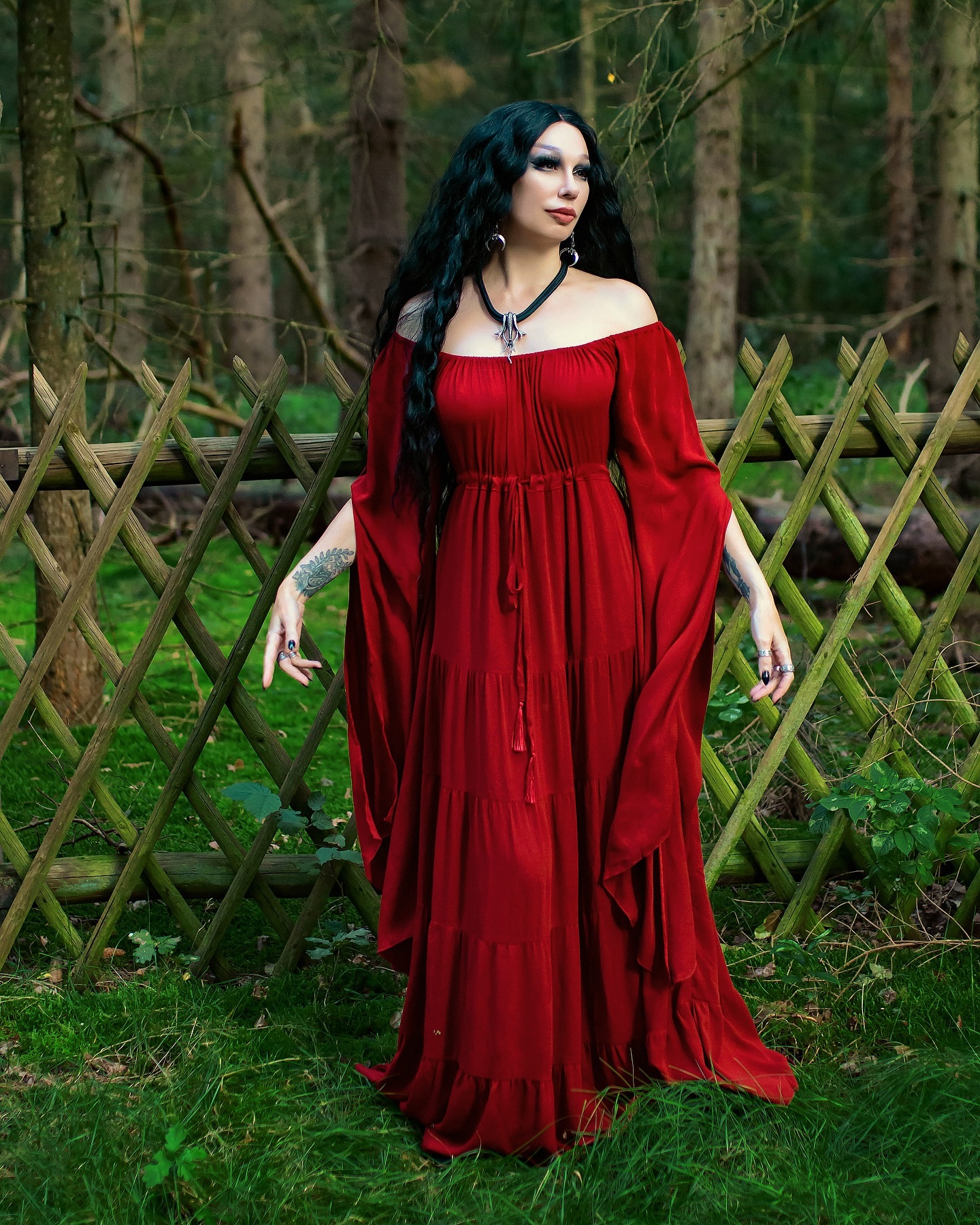 a fullbody portrait of a woman in a long, flowing, tiered red offtheshoulder dress. she has long dark hair, and is wearing a dark necklace. she is standing in a forest setting next to a wooden fence, with lush green grass around her. the overall mood is dark, mystical, and ethereal. the dress has a bohemian or medieval style, and the model's pose and attire suggest a fantasy or gothic aesthetic.