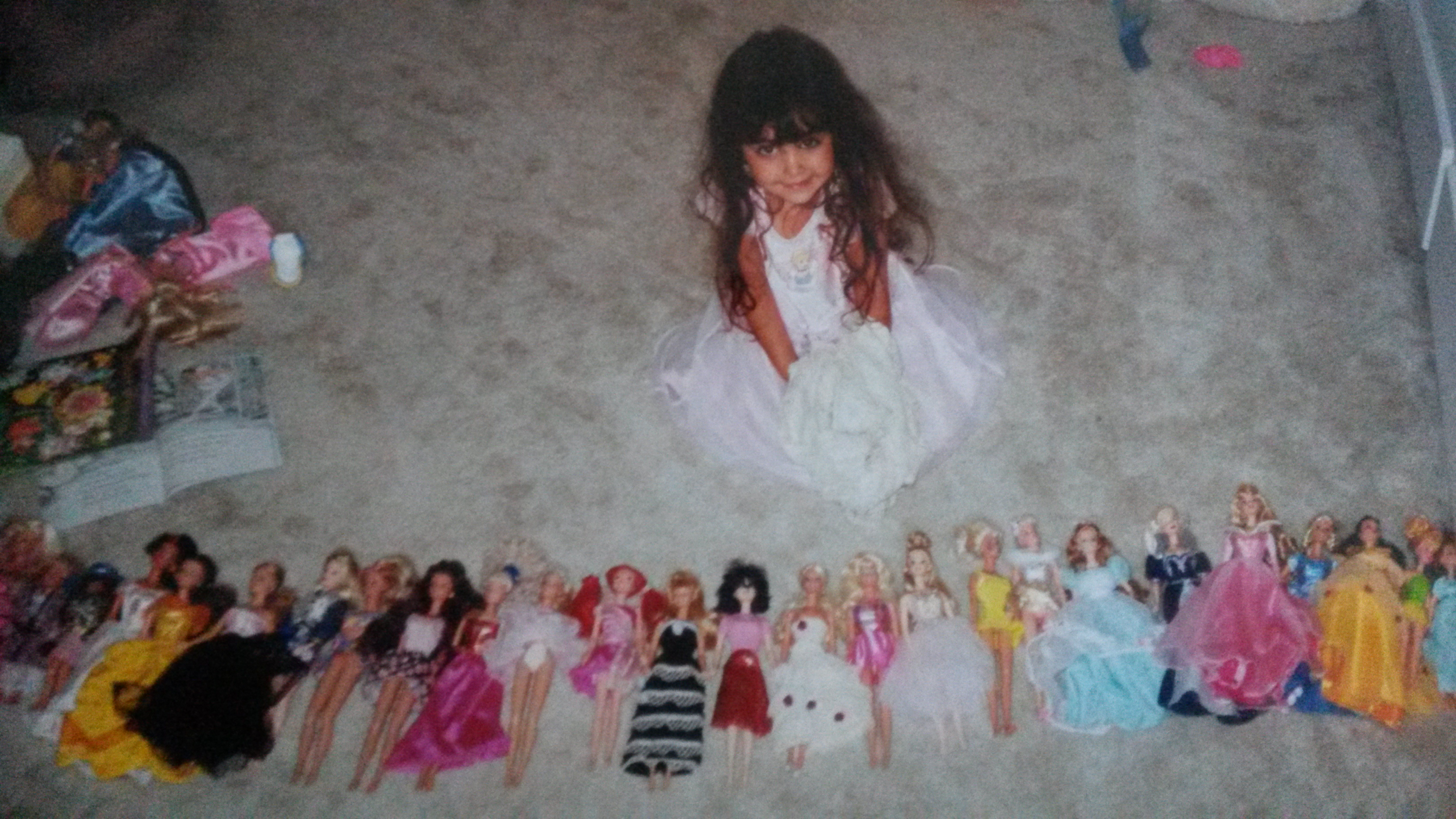 a young girl, around 35 years old, with dark brown hair sits on a carpeted floor surrounded by a large collection of barbie dolls. she is wearing a white dress. the dolls are arranged in a row in front of her, each wearing a variety of colorful dresses.  some other items are visible in the background, including what appear to be other toys and books. the overall impression is one of a childhood memory, perhaps a photo taken in the 1980s or 1990s.