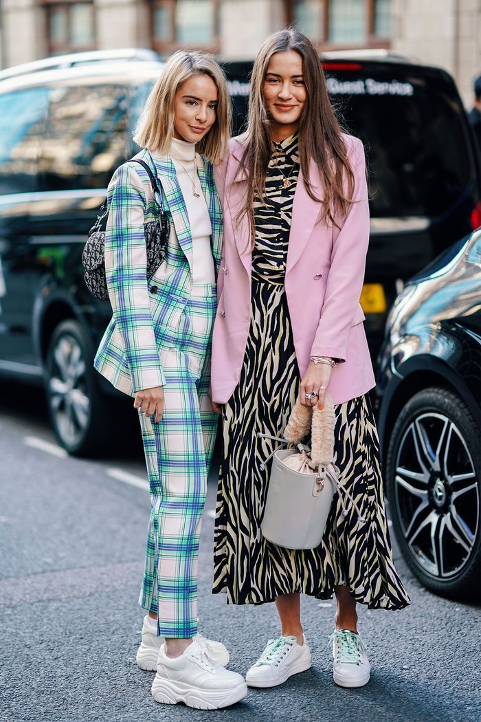 two stylishly dressed women are standing on a city street. one is wearing a green and white plaid pantsuit with white chunky sneakers. the other is wearing a pink blazer over a zebraprint midi dress and white sneakers. they both carry handbags.  the background includes cars and city buildings. the overall aesthetic is one of contemporary street style fashion.