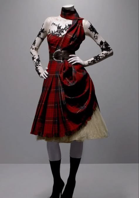 a fashion model is wearing a red and black plaid kilt with a black lace top and a black belt with a silver buckle. the kilt is draped over one leg, creating a dramatic silhouette. the model is wearing black tights and highheeled boots. the model is standing on a gray background. this is a runway look.