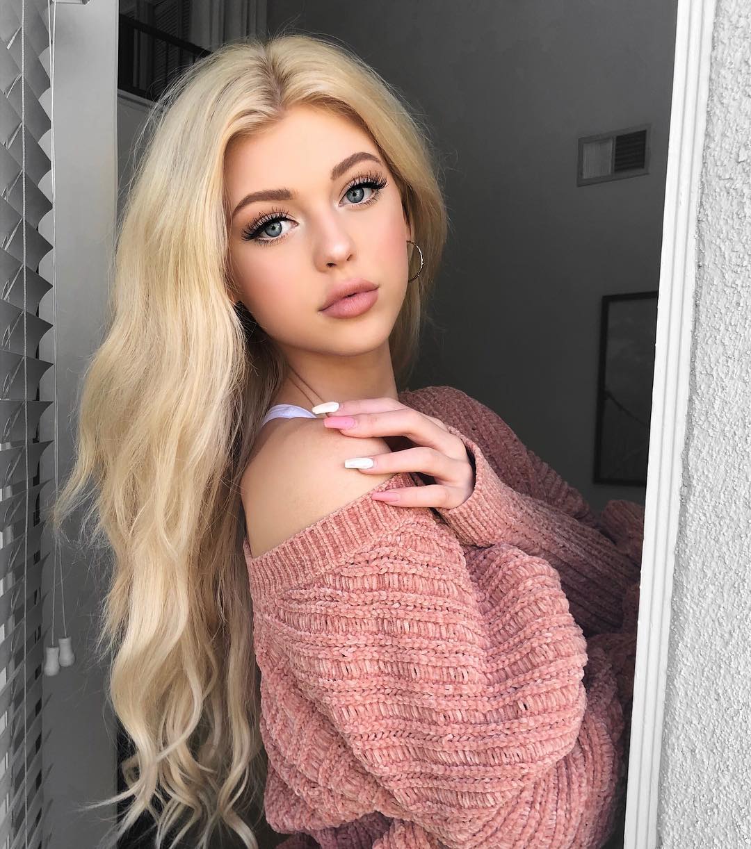a woman with long blonde hair and blue eyes wearing a pink offshoulder sweater is looking at the camera indoors, positioned near a window. she is wearing hoop earrings and has long painted nails.