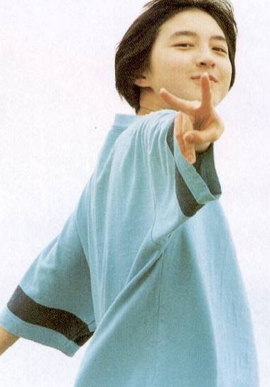 a young asian boy is wearing a light blue shirt with a black stripe on each sleeve. he is smiling and making a peace sign with his hand. the background is white.
