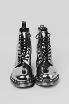 a pair of silver dr. martens ankle boots are placed side by side on a light background. they are made of shiny leather and feature black laces. the boots are shown from the front.