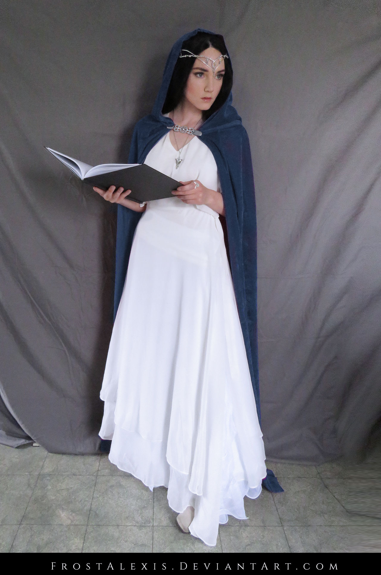 a fullbody shot of a woman dressed in an elf costume. she's wearing a long white dress under a dark blue hooded cloak. a silver tiara rests on her forehead. she holds a book in her hands. the background is a plain grey backdrop. the image seems to be a cosplay or character portrayal.
