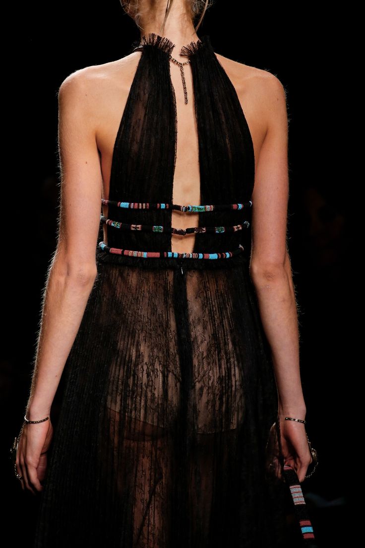 a woman wearing a black sheer lace dress with a low cut back on the runway during a fashion show. the dress has beaded straps and an open back, and she is accessorized with silver jewelry.