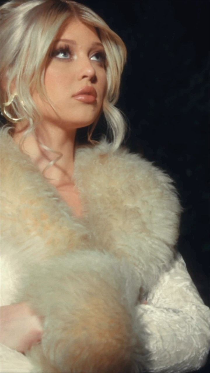 a portrait of a woman with blonde hair wearing a fur collar and earrings, looking up. the background is dark.