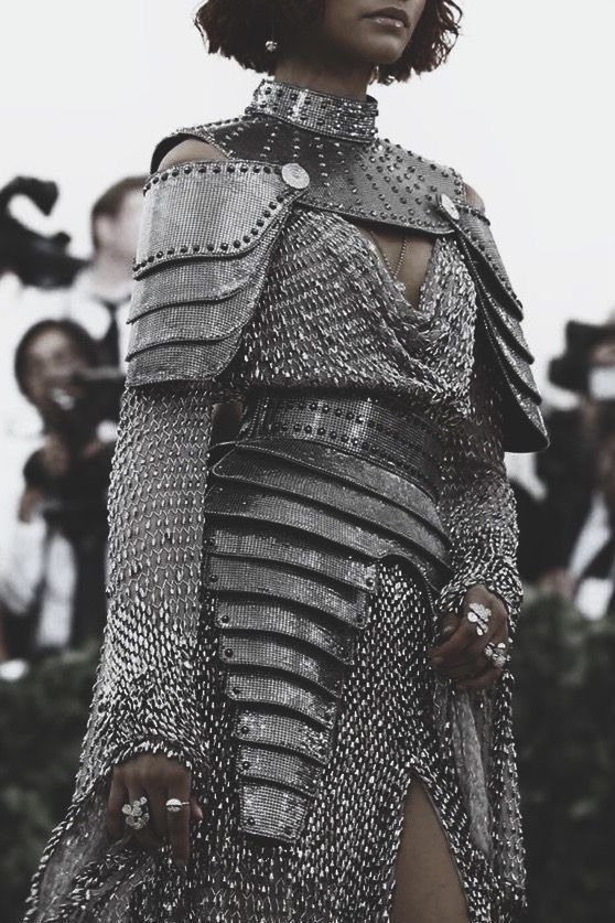 the image shows a woman wearing a striking silver chainmail dress that resembles armor. the dress features a high neck, shoulder pauldrons, and a segmented skirt. the intricate detailing, including studs and metallic sheen, creates a glamorous yet edgy look. the background is blurred, suggesting an outdoor event with onlookers. the overall aesthetic blends fantasy and high fashion.