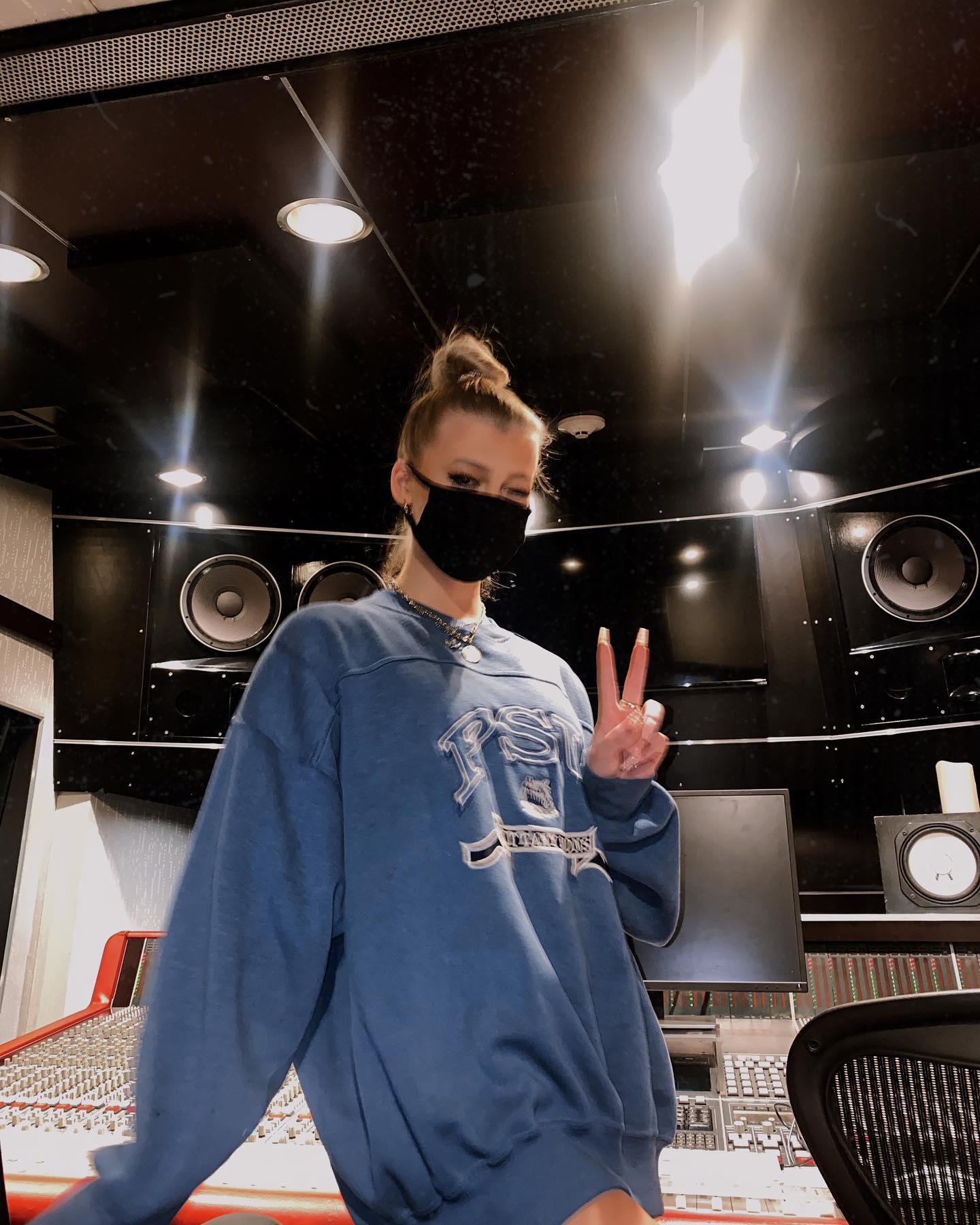 a woman wearing a blue sweatshirt and a face mask makes a peace sign while standing in a recording studio. the studio has various audio equipment, speakers, a monitor, and a chair. the woman has her hair in a messy bun.