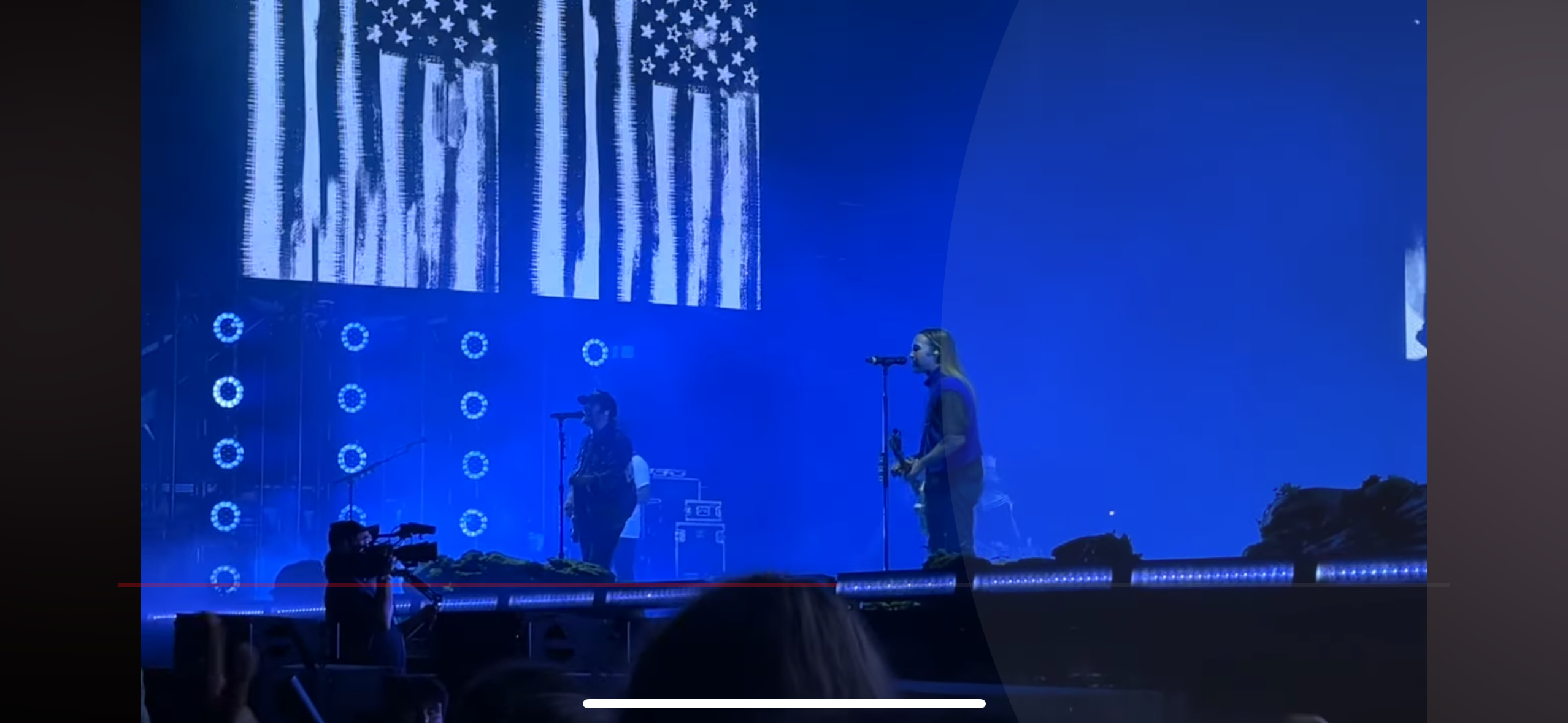 a live music concert performance on a stage with a band playing. there is a singer with a microphone and a guitar player. the stage is lit with blue lights and there is an american flag in the background. the crowd is in the foreground and you can see the backs of people's heads. the concert is happening at night.