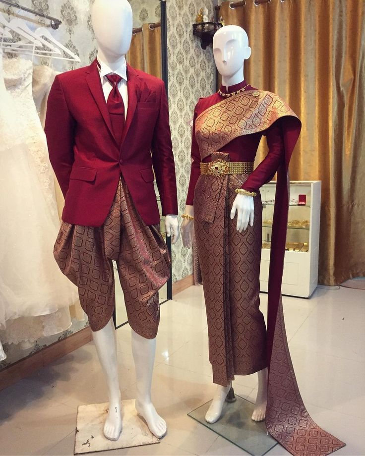 a bride and groom are wearing traditional thai wedding attire. the groom is wearing a red suit and the bride is wearing a red dress with gold trim and a long train. they are standing in front of a white backdrop and there is a wedding dress hanging on the left.