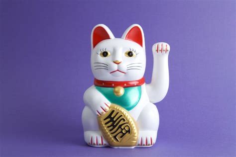 a closeup shot of a white manekineko (beckoning cat) figurine against a solid purple background. the cat is depicted with a red collar, teal bib, and is holding a gold coin in its paw. one paw is raised in a beckoning gesture.  the figurine is detailed and appears to be made of ceramic or plastic. the image is welllit and professionally photographed.